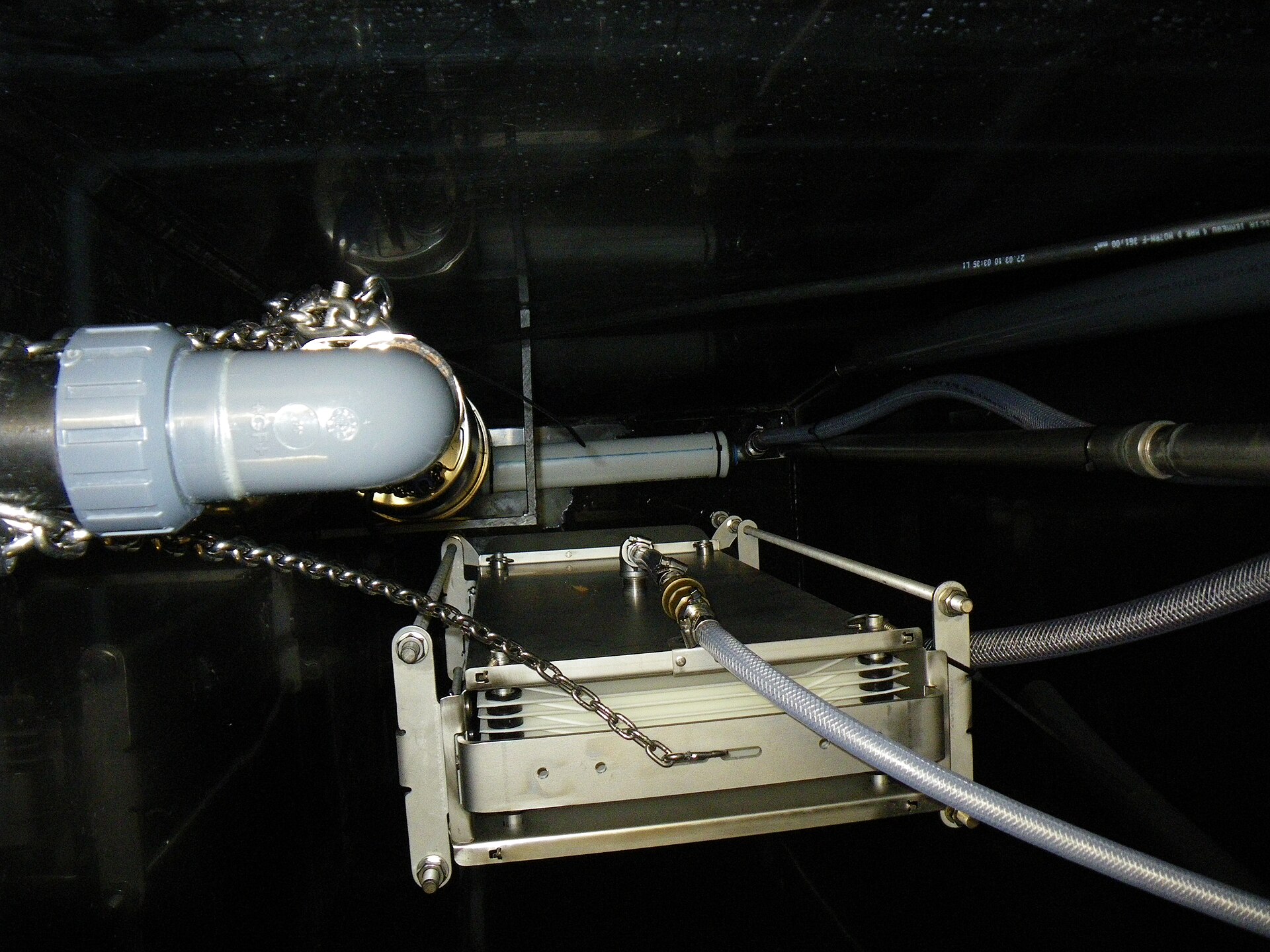 A steel membrane filtration module hanging in a dark tank, fed by hoses and held by a chain