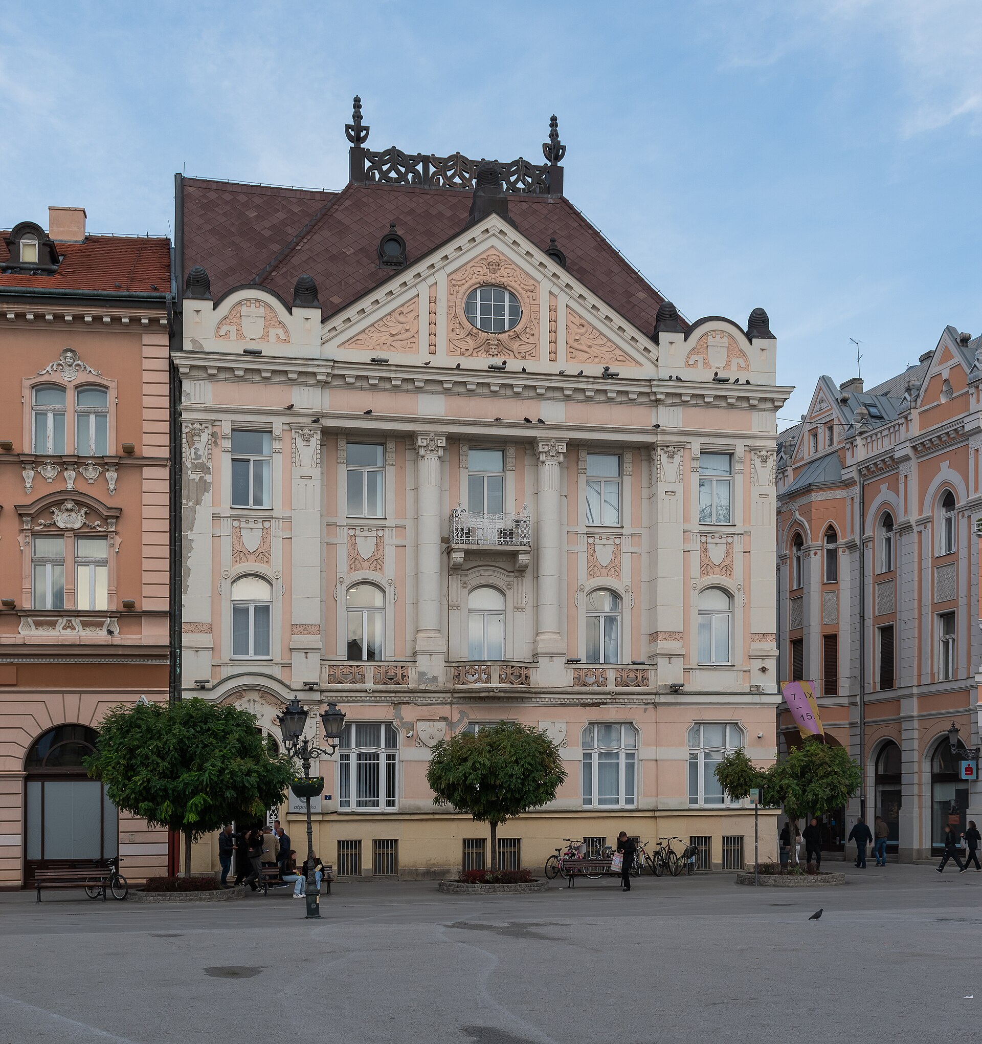 A pale pink Secession building with an ornate gable on Novi Sad’s main square, Trg slobode, with trees and people in front