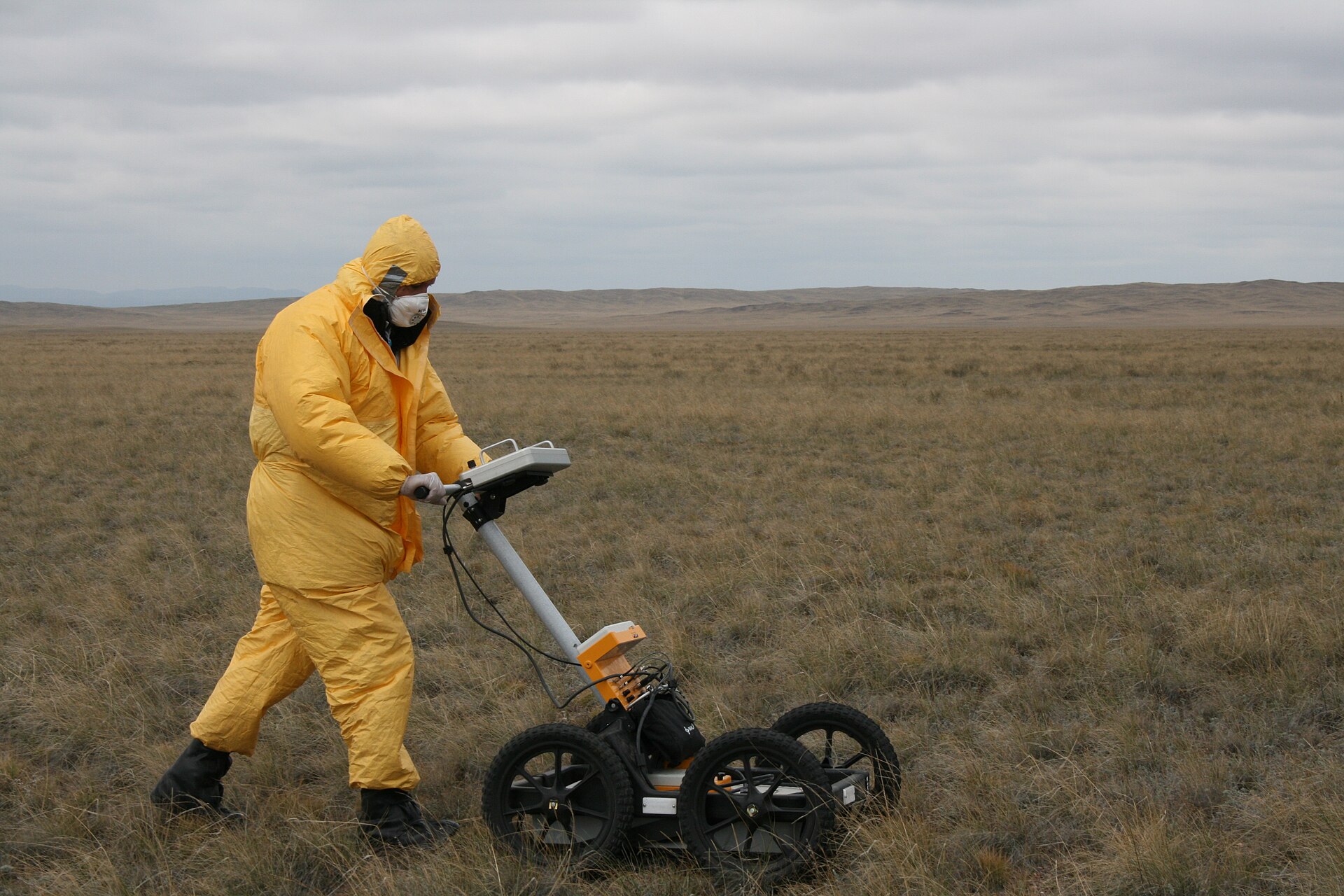 A surveyor in a yellow protective suit pushes a ground-penetrating radar cart across open grassland